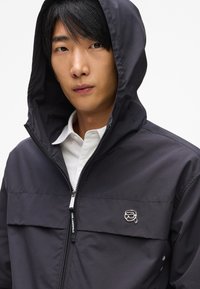 Sötétkék kapucnis dzseki sima anyagból, elöl cipzárral, mellzsebbel és logós díszítéssel. Fehér galléros ing felett viselve.