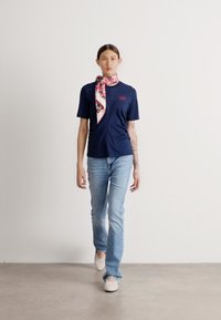 T-shirt navy a maniche corte con una sciarpa a fantasia floreale. Abbinato a jeans azzurri e scarpe slip-on bianche. Sfondo semplice.