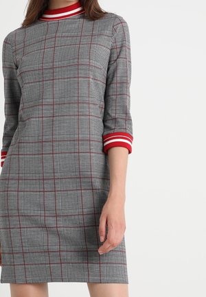 Kobieta ubrana w szarą sukienkę w kratę z czerwono-białym paskowanym kołnierzykiem i mankietami na rękawach, stojąca na jednolitym białym tle.