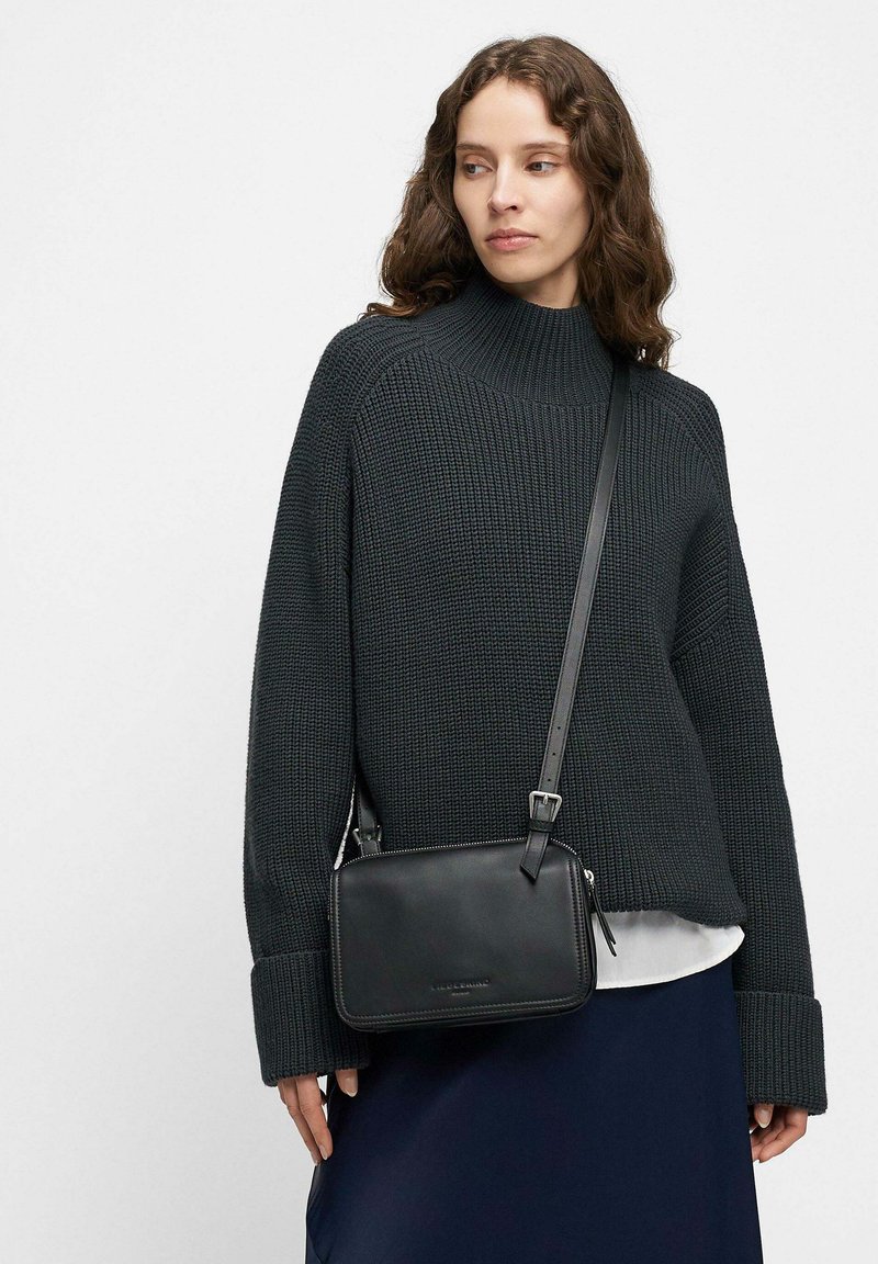 Borsa a tracolla in pelle nera con chiusura a zip, tracolla regolabile, forma rettangolare e design minimalista. Indossata con un maglione a trama.