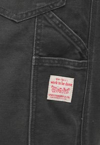 Τσέπη ρούχου από μαύρο ντένιμ με λευκή ετικέτα Levi Strauss & Co., όπου αναγράφεται το μήνυμα «όταν υπ άρχει δουλειά να γίνει» με κόκκινο κείμενο.