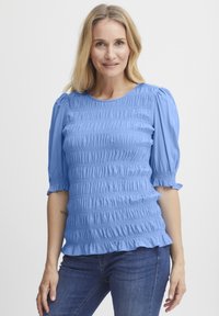 Lyseblå smockbluse med korte puffærmer og en rynket kant. Lavet af blødt, tekstureret stof. Bæres med mørkeblå jeans.