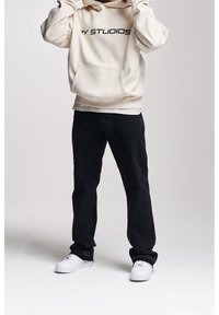 Sudadera crema con texto negro, bolsillo delantero, combinada con jeans negros y zapatillas blancas, mostrando un estilo atlético casual.