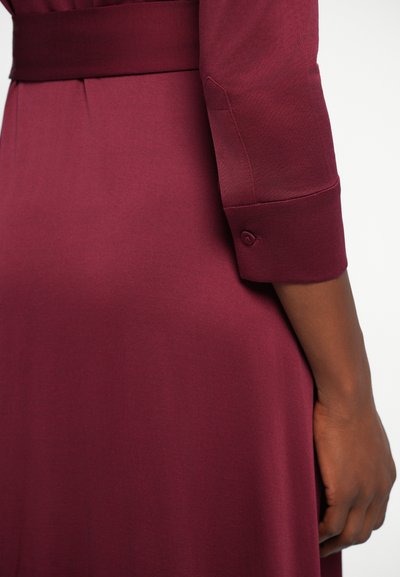 Burgundska haljina glatke teksture, dugih rukava i s detaljem preklopnog manžeta. Ima usku liniju struka s odgovarajućom trakicom.