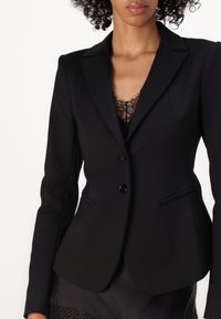 Černý blazer vyrobený z hladkého materiálu, s klopovým límcem, dvěma knoflíky a bočními kapsami, nošený přes černý krajkový top.