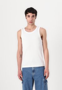 Witte katoenen tanktop met een ronde halslijn, voorzien van een geribbelde textuur. Het model draagt blauwe denim cargo shorts.