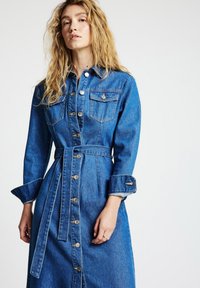 Denimkleid in mittlerem Blau mit langen Ärmeln, knöpfbarem Vorderteil, Brusttaschen und einem taillierten Gürtel. Verfügt über einen geraden Schnitt und gekrempelte Ärmel.
