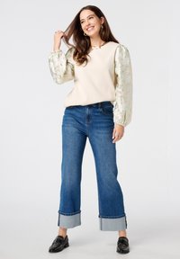 Haut crème avec des manches bouffantes texturées et à motif ; associé à un jean large bleu foncé et à des mocassins noirs à clous.