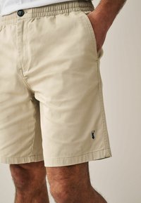 Pantaloni corti beige realizzati in tessuto leggero, con vita elasticizzata e bottone, tasche laterali e un piccolo logo blu navy.