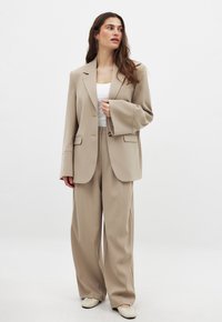 Bež zmogljivo oblikovan sako z urejeno jakno in širokimi hlačami, v kombinaciji z belo majico na tanke naramnice in svetlim kremnim natikačem.