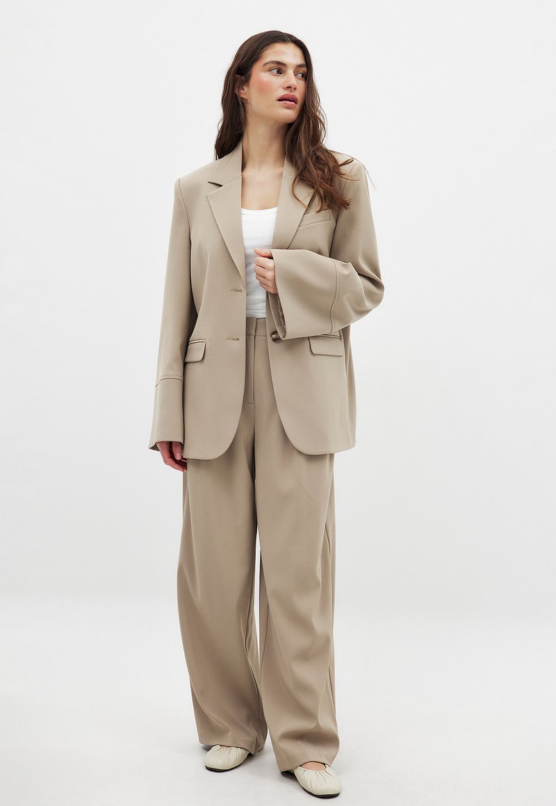 Bež zmogljivo oblikovan sako z urejeno jakno in širokimi hlačami, v kombinaciji z belo majico na tanke naramnice in svetlim kremnim natikačem.