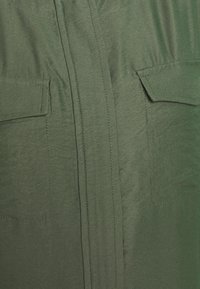 Zbliżenie na oliwkowozielony materiał z dwiema naszytymi kieszeniami, z jedną pokrytą klapką oraz widocznymi pionowymi przeszyciami.