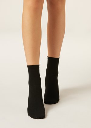 Nohy v jednoduchých černých kotníkových ponožkách stojící na světlém povrchu na neutrálním pozadí.