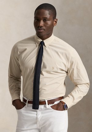 Vyras dėvi smėlio spalvos langeliais margintą marškinių, juodą tekstūruotą kaklaraištį, baltas kelnes, rudą diržą ir mėlynu ciferblatu laikrodį, stovintis su rankomis kišenėse.