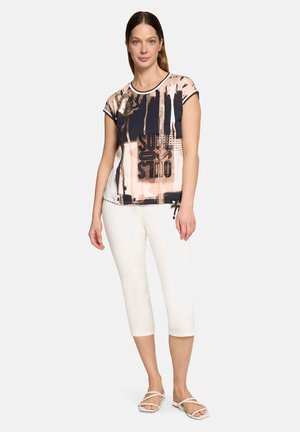 Frau trägt ein kurzärmeliges Oberteil mit abstraktem Muster und Text sowie weiße, knöchellange Hosen und steht mit einer Hand entspannt an ihrer Seite.