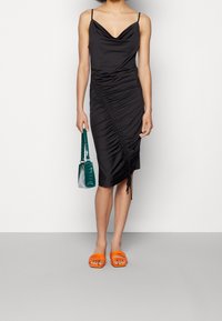 Žena v černých midi šatech s rouchou a tenkými ramínky, držící zelenou kabelku a obutá v jasně oranžových sandálech s otevřenou špičkou, stojící proti bílé zdi.