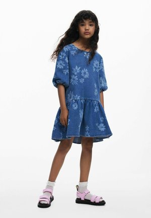 Jeanskleid mit blauen Blumenmustern, Puffärmeln und einem mehrstufigen Rock. Kombiniert mit pinken Sandalen und weißen Socken.