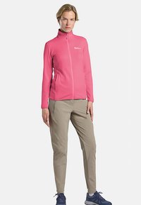 Giacca in pile rosa acceso con cerniera integrale e tasche laterali, abbinata a pantaloni beige e scarpe da ginnastica blu.