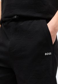 Μαύροι σορτς με υφή, με πλαϊνή τσέπη και λευκό λογότυπο "BOSS", με το χέρι εν μέρει μέσα στην τσέπη και ελαστική μέση με κορδόνι ορατό.