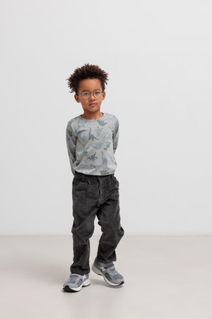 Child with curly hair and glasses stands with hands behind back, wearing a dinosaur-patterned shirt and corduroy pants in a minimalist setting.