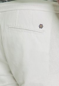 Witte katoen broek met een enkele achterzak, voorzien van een hexagonale gouden en zwarte accentbadge, die de gladde textuur en de op maat gemaakte uitstraling benadrukt.