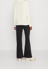 Person klädd i en krämfärgad tröja, svarta utsvängda jeans och kraftiga off-white stövlar, stående mot en plain vit bakgrund.