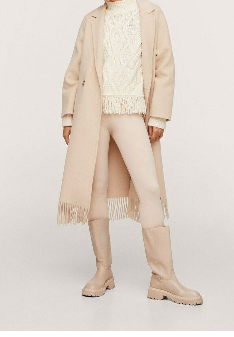 Personne portant un long manteau beige à franges, un pull en maille torsadée crème, des leggings beige clair et des bottes hautes beige, debout devant un fond uni.