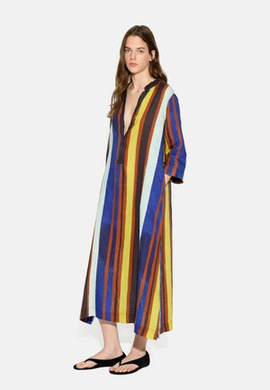 Kobieta stojąca w długiej, luźnej sukience z pionowymi niebieskimi, żółtymi, brązowymi i pomarańczowymi paskami oraz czarnych sandałach typu japonki.