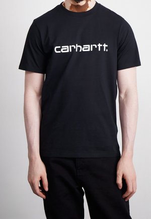 Personne portant un t-shirt noir Carhartt avec un logo blanc, montrant les bras et le torse sur un fond clair uni.