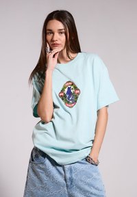 Camiseta de algodón de color azul claro con mangas cortas, que presenta un gráfico central de una criatura marina púrpura rodeada de detalles verdes.