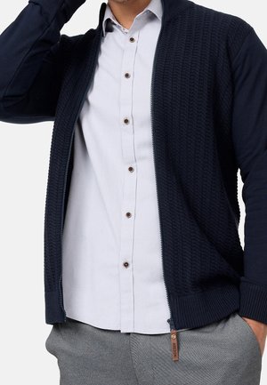 Homme portant un cardigan zippé texturé bleu marine sur une chemise blanche à boutons, avec un pantalon gris, main dans la poche et bras levé.