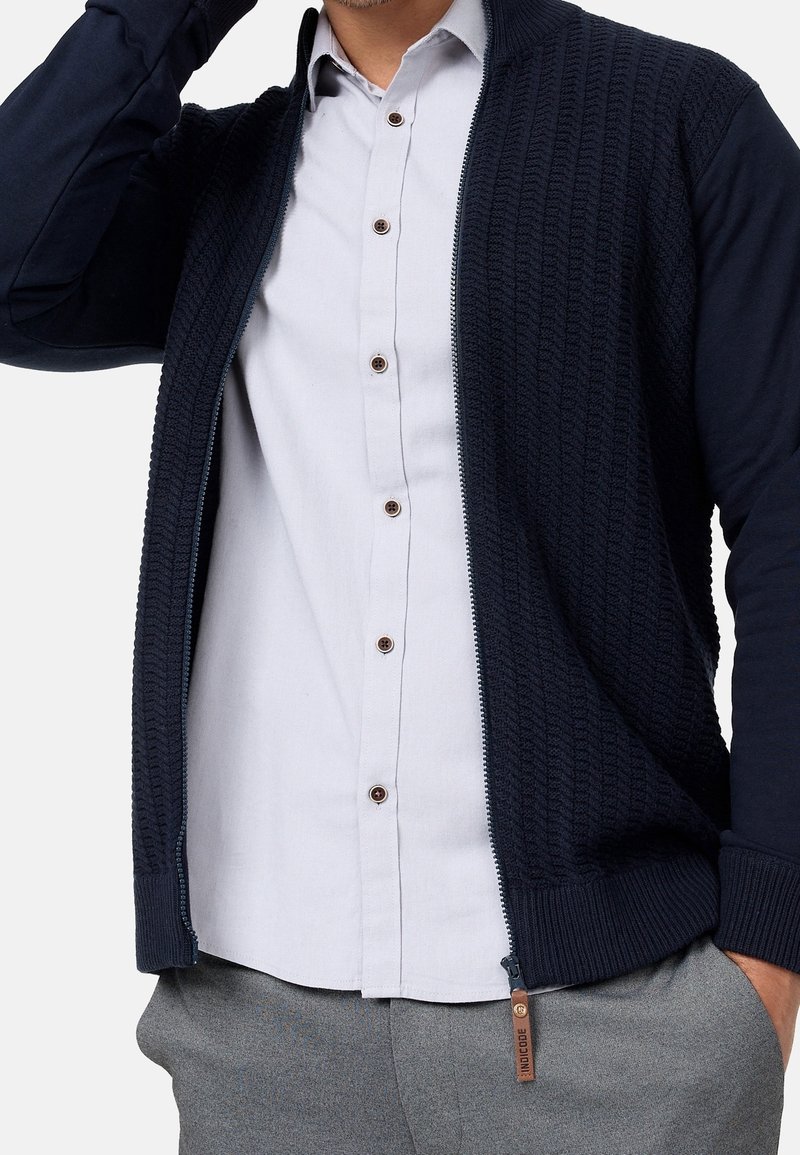 Homme portant un cardigan zippé texturé bleu marine sur une chemise blanche à boutons, avec un pantalon gris, main dans la poche et bras levé.
