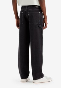 Breda svarta denimjeans med två bakfickor, stygnaccent och en subtil sliten textur, matchade med vita sneakers.