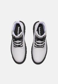 Bottines en cuir blanc avec semelle en caoutchouc noir, dotées de lacets noirs et d'un col rembourré. Logo Timberland visible sur la semelle intérieure.