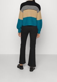 Frau in einem farbblockierten Pullover mit schwarzen, beigen und petrolfarbenen Abschnitten, schwarzen Schlaghosen und schwarzen Loafers, die vor einem weißen Hintergrund steht.