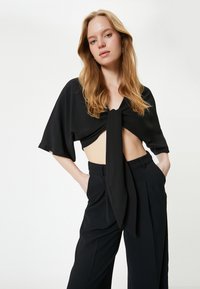 Sort cropped top med knyttet foran, korte ærmer og løstsiddende fit. Stylet med højtaljede, skræddersyede sorte bukser med sidelommer.
