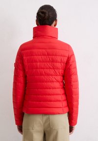 Osoba z włosami związanymi w kok, ubrana w jasnoczerwoną pikowaną kurtkę, widziana od tyłu, stojąca na tle jednolitej jasnej ściany.