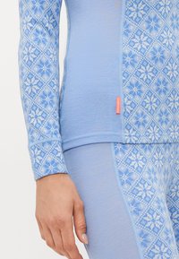 Nærbilde av en person som har på seg en lyseblå mønstret langermet topp og leggings med snøfnuggdesign og et lite oransje merketagg.