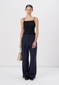 Schwarzes geripptes Tanktop, weite marineblaue Hose und schwarze Schuhe. Hält eine cremefarbene Handtasche mit Kettenriemen. Einfaches, minimalistisches Design.