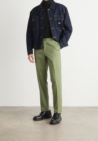 Giacca di jeans con due tasche sul petto, blu scuro; dolcevita nero; pantaloni verde chiaro; scarpe nere. Design semplice e vestibilità sartoriale.