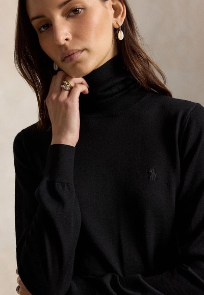 Femme portant un pull à col roulé noir avec un petit logo brodé, des boucles d'oreilles en forme de coquillage et plusieurs bagues aux doigts touchant son menton.