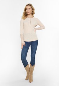 Beige hosszú ujjú felső gombokkal, csipke díszítéssel az ujjakon, sötétkék farmernadrággal és bézs cowboy csizmával, díszített varrással párosítva.