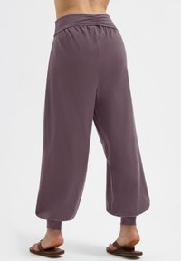 Person, der weite, hoch taillierte mauvefarbene Hosen mit gerafften Bündchen trägt und barfuß auf weißem Hintergrund steht, Rückansicht.