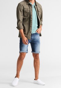 Camisa de botón verde oliva sobre un polo azul claro, combinada con pantalones cortos de mezclilla enrollados y zapatos de tela blancos. Atuendo simple y casual.