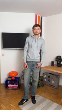 Grå hettegenser med glidelås, med en liten gul logo. Lyseblå jeans, svarte sko med hvite striper, tatoveringer synlige på hendene.