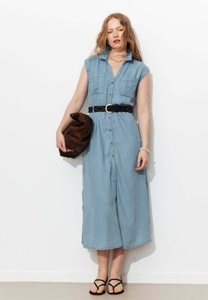 Femme portant une robe en jean sans manches boutonnée avec une ceinture noire, tenant une pochette marron à motifs, et portant des sandales noires, debout contre un mur blanc.