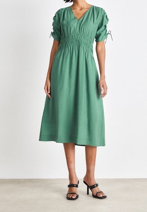 Kobieta w zielonej sukience midi z bufkami na rękawach i marszczonym pasem, w połączeniu z czarnymi sandałami na obcasie, stojąca na jednolitym tle.