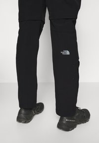 Jambes portant un pantalon d'extérieur noir avec le logo The North Face et des chaussures de trail Mammut noires sur un fond blanc uni.
