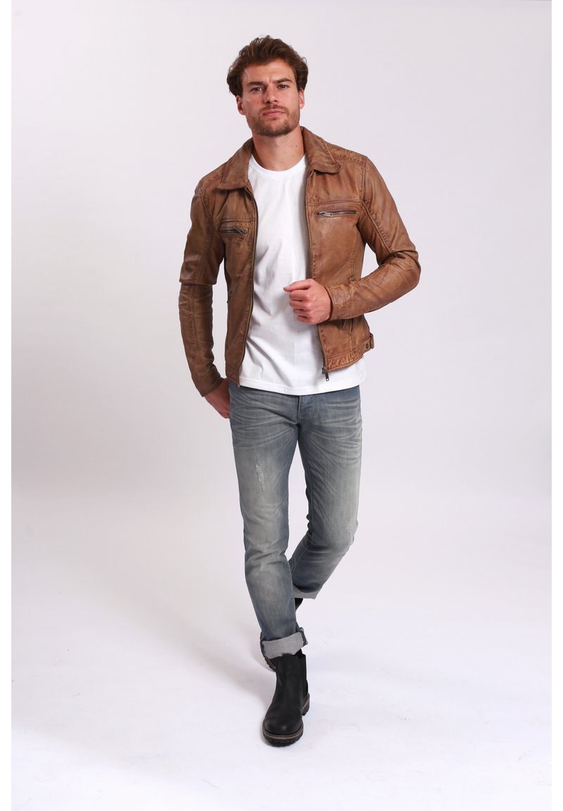 Braune Lederjacke mit Reißverschluss und Taschen, kombiniert mit einem weißen T-Shirt und hellblauen, stark verwaschenen Jeans. Schwarze Ankle Boots runden das Outfit ab.