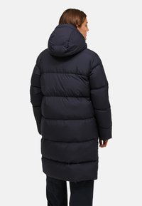Kvinna i lång svart pufferjakka med stor huva, sedd bakifrån mot en neutral bakgrund.